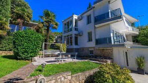 A modern two-story house with a stone foundation, a balcony on the upper floor, and a small garden area with a bench and palm trees. - Holiday apartment with air conditioning (Crikvenica)