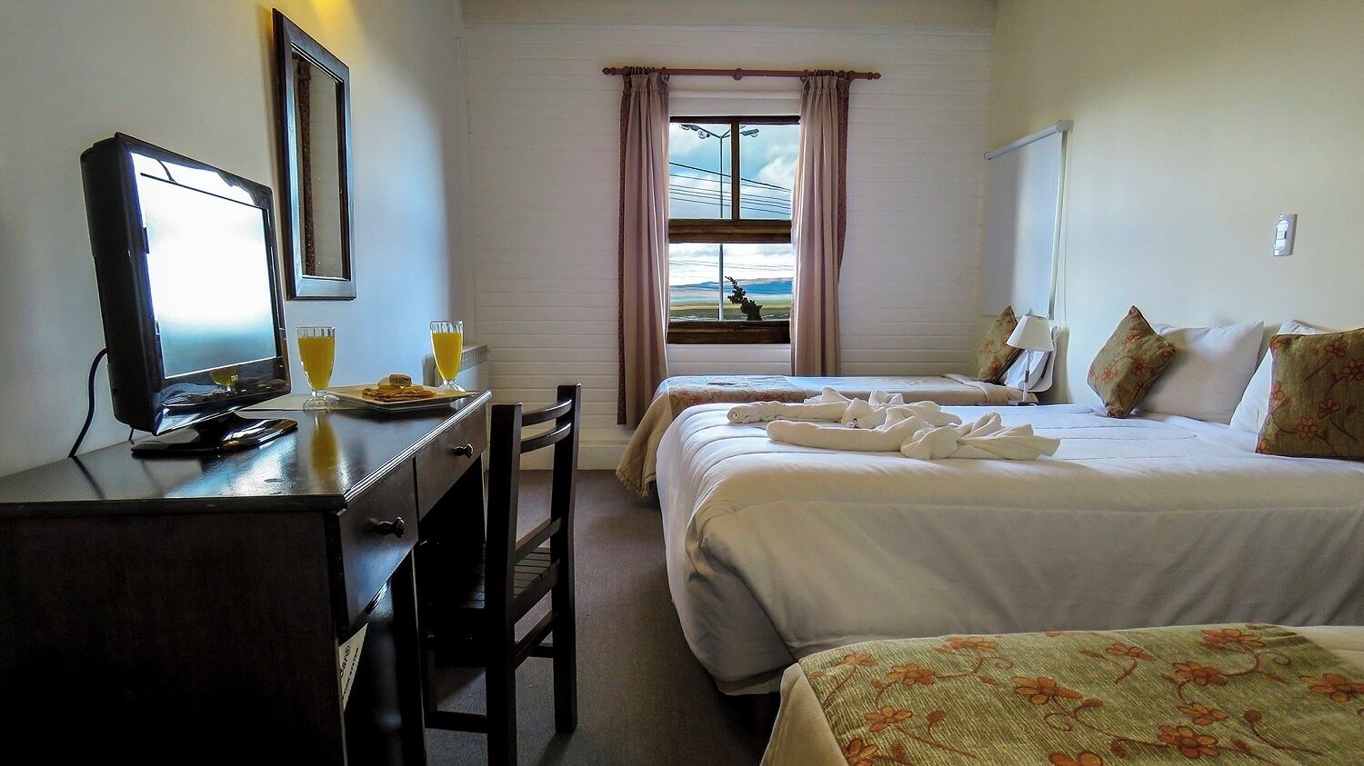 A hotel room with a desk, two beds, a window with curtains, and a view of the outdoors.