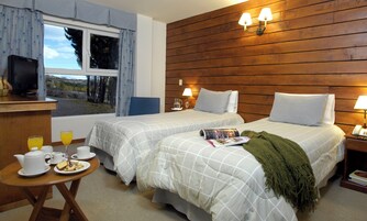 Habitación de hotel con dos camas, cabecera de madera, televisión, una mesa con alimentos para el desayuno y vista a árboles por la ventana.