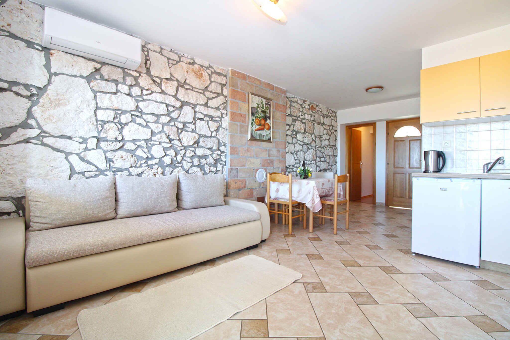 A living room with a stone wall, a sofa, a dining table with chairs, and a kitchen area with cabinets and a refrigerator.