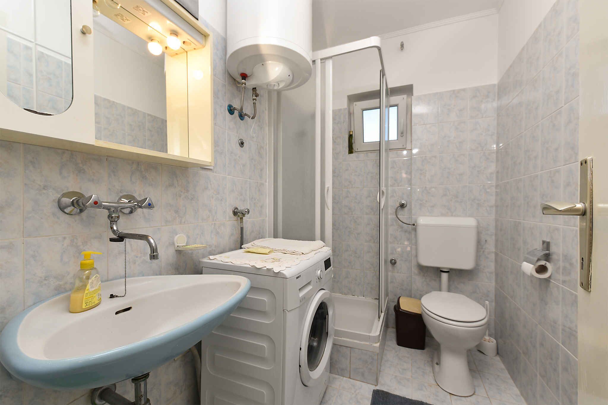 A compact bathroom with a sink, washing machine, toilet, and shower enclosure.