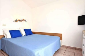 Un lit simple avec une couvre-lit bleue et deux oreillers, un téléviseur fixé au mur et un sol carrelé.