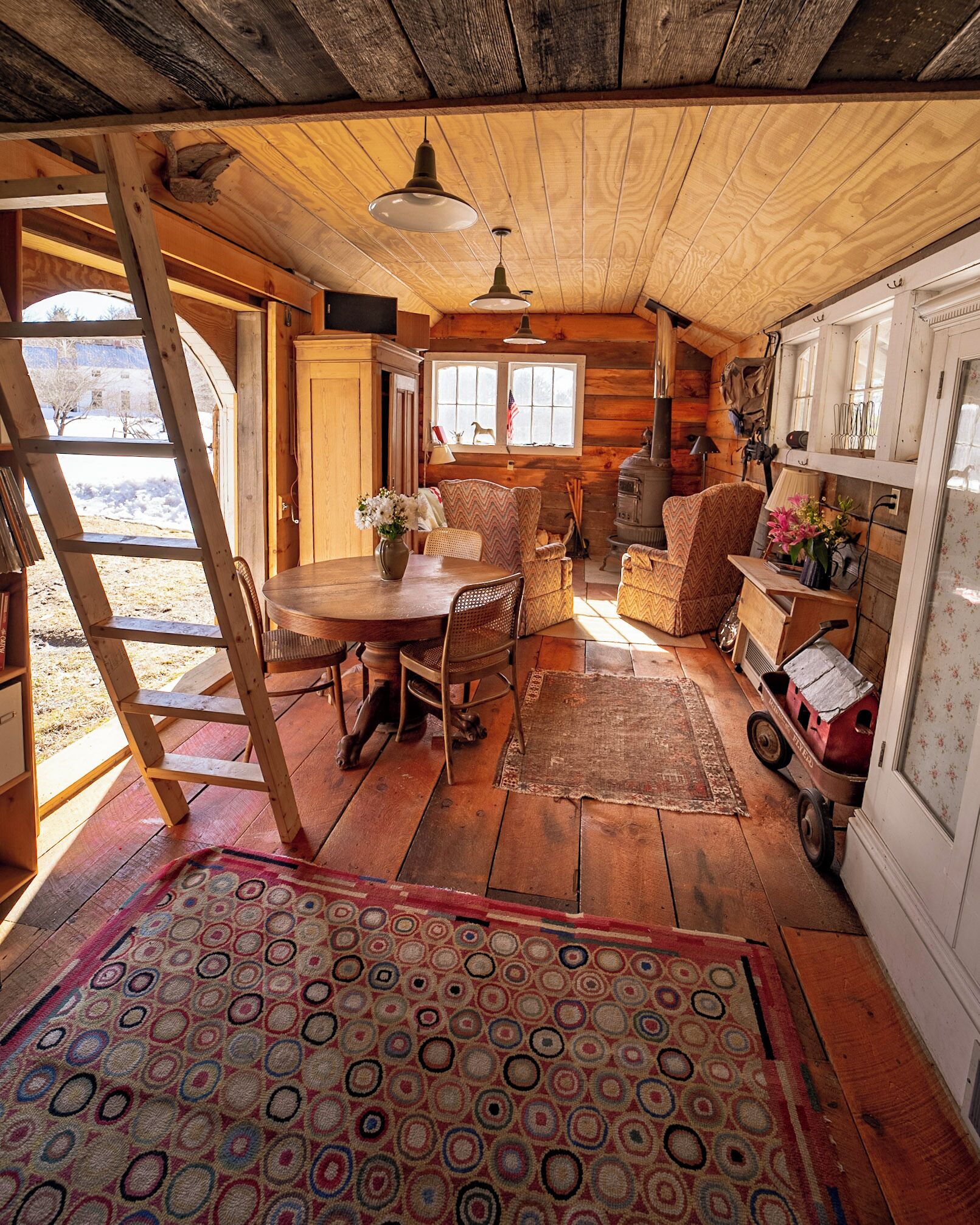 Interno accogliente di una baita rustica con un tavolo da pranzo in legno, sedie, un tappeto decorato e un vecchio stufa.