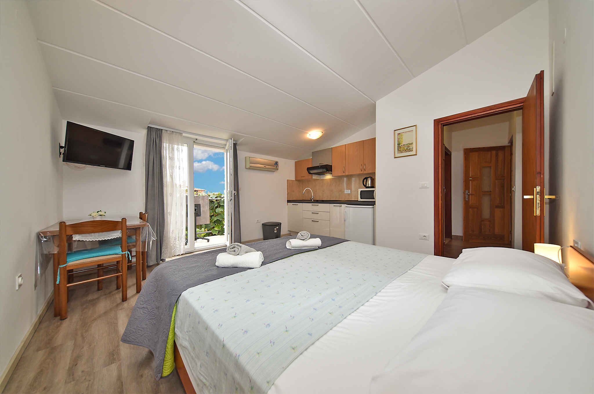 Quarto de hotel com cama, escrivaninha, televisão e cozinha compacta.