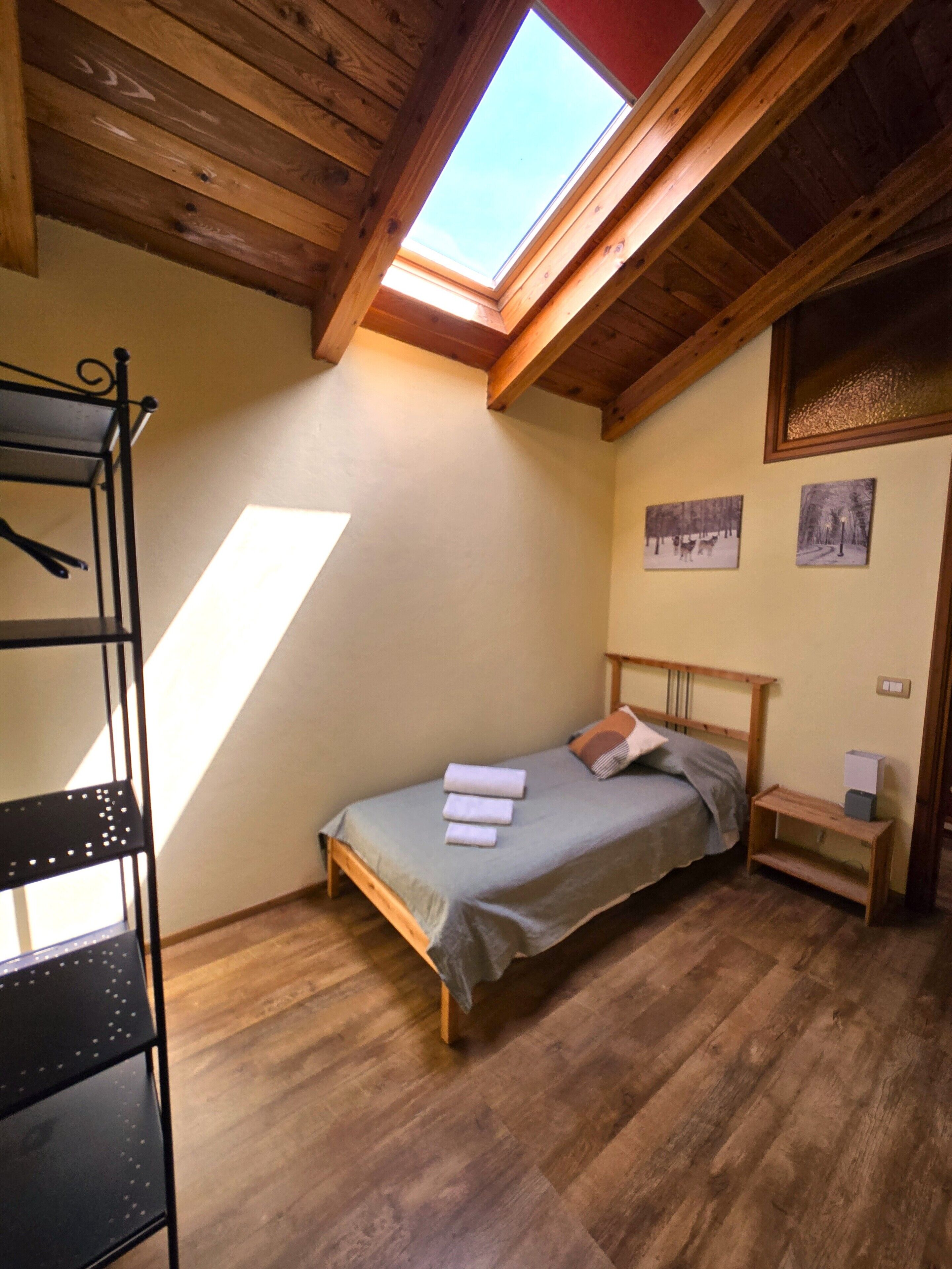 Ein Schlafzimmer mit Dachfenster, Holzbalken, einem Bett mit Kissen, einem Nachttisch und einem Regal.