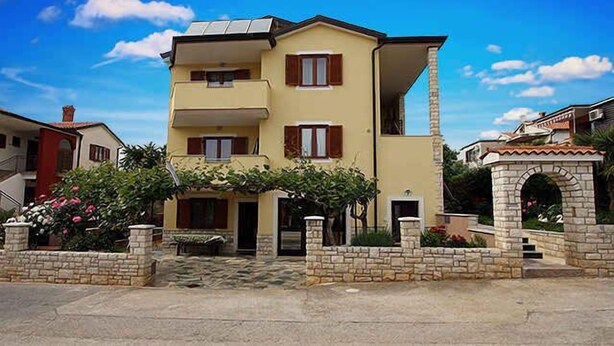 A two-story building with a balcony on the upper floor, a stone entrance, and a garden area with flowers.