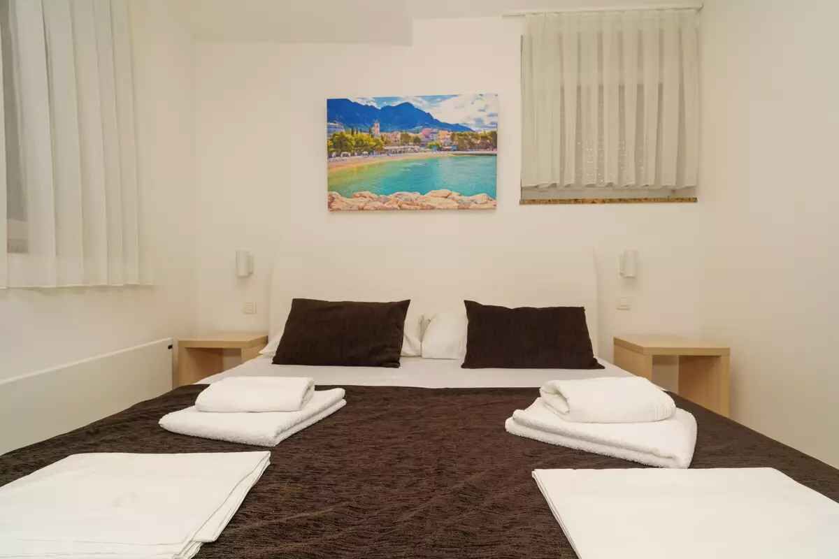 A hotel room with a bed, two pillows, a painting of a coastal scene, and a window with curtains.