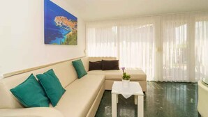 A modern living room with a beige sofa, teal cushions, a small white table with a potted plant, and a large window with sheer curtains. - Holiday apartment with air conditioning and barbecue facilities (Crikvenica)