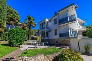 Une maison moderne à deux étages, avec des fondations en pierre, un balcon à l’étage supérieur et un petit jardin agrémenté d’un banc et de palmiers.