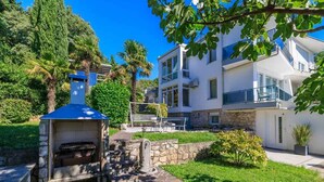 A modern building with a stone barbecue area, a stone wall, and a well-maintained garden with palm trees. - Holiday apartment with air conditioning and barbecue facilities (Crikvenica)