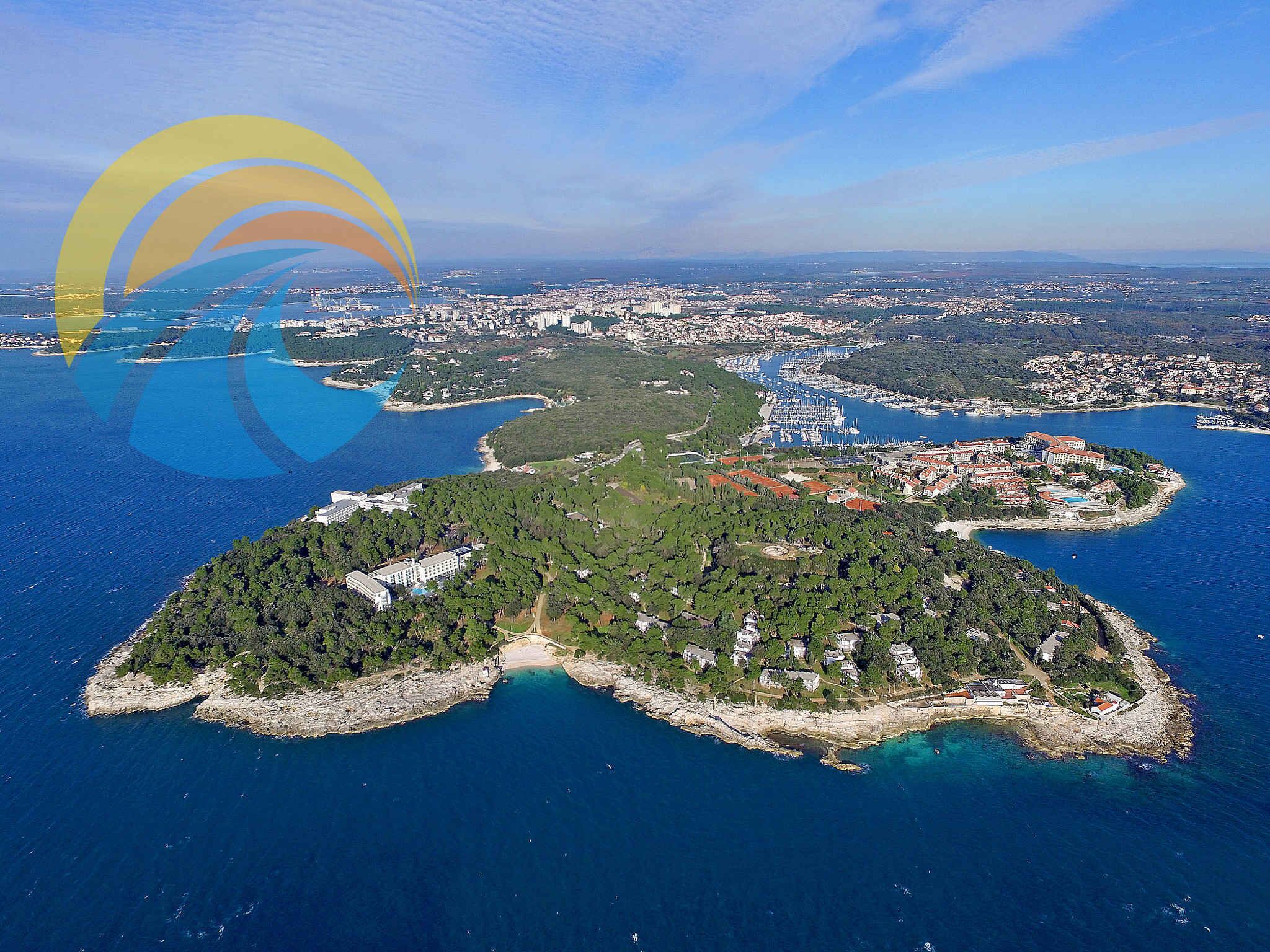 An aerial view of a coastal town with a marina, green hills, and clear blue waters.