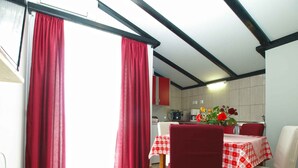 Un coin repas avec une table dressée pour deux personnes, des rideaux rouges et une cuisine en arrière-plan.
