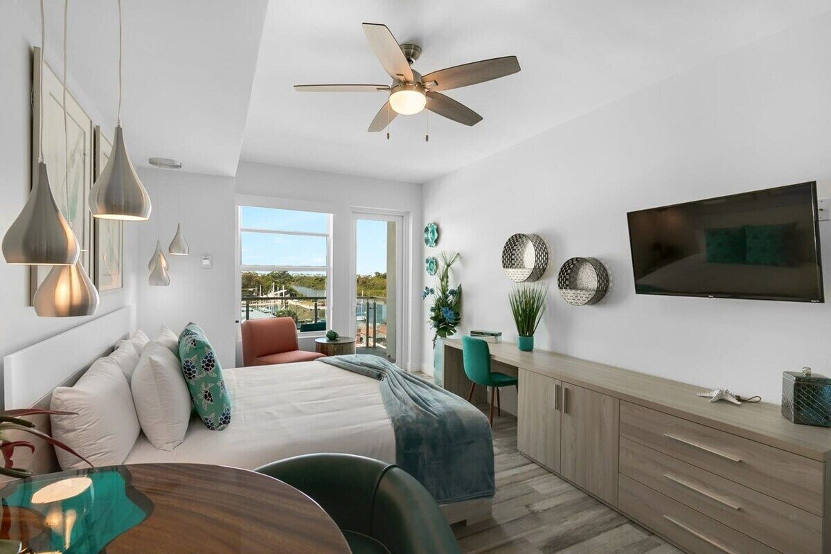 A modern hotel room with a bed, desk, and TV. There are pendant lights, a ceiling fan, and a view of the outdoors.