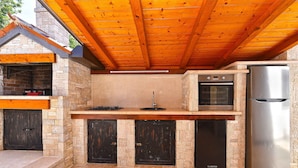 Una cocina al aire libre con pared de piedra, techo de madera y electrodomésticos integrados, incluyendo estufa, horno y refrigerador.