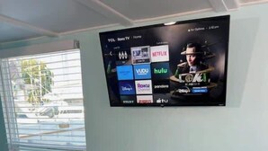 Una habitación con una mesa y sillas color turquesa, un televisor de pantalla plana montado en la pared que muestra varios servicios de streaming, y una ventana con persianas.