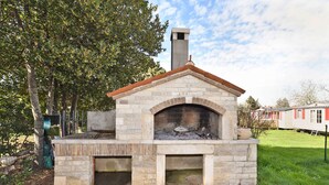 A stone-built outdoor pizza oven with a chimney, surrounded by a stone patio and a grassy area with trees. - Holiday apartment with garden and air conditioning (Medulin)