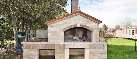A stone-built outdoor pizza oven with a chimney, surrounded by a stone patio and a grassy area with trees.