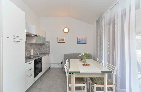 Eine kompakte Küche mit weißen Schränken, einem Einbauofen und einer gefliesten Spritzschutzleiste. Es gibt einen Essbereich mit Tisch und Stühlen, sowie ein Fenster mit durchsichtigen Vorhängen.