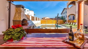 Eine Terrasse mit Kamin aus Stein, einem Tisch mit Streifen-Tischdecke, einer Weinflasche und einem Blumentopf.