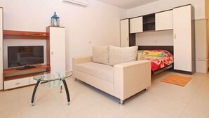 A compact living space with a sofa, a glass coffee table, a TV on a wooden stand, and a wardrobe with a bed underneath.