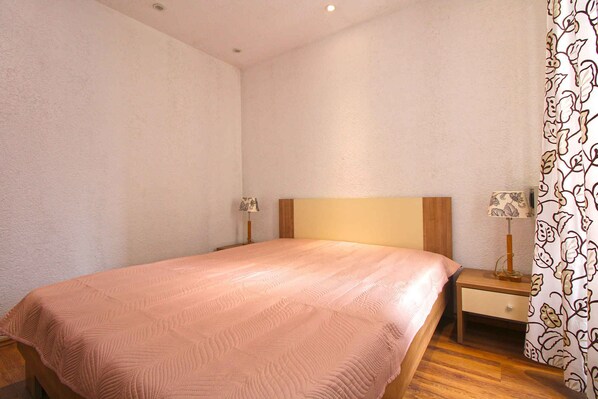 Una cama bien hecha con una colcha rosa, una mesita de noche de madera con una lámpara y una cortina estampada.