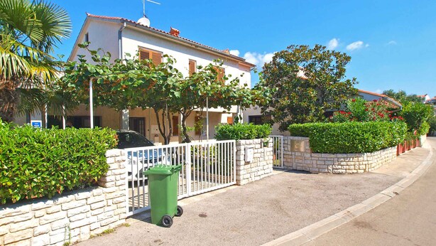 A residential street with a white house, a green trash bin, and a stone wall. - Holiday apartment with air conditioning (Pješćana Uvala)