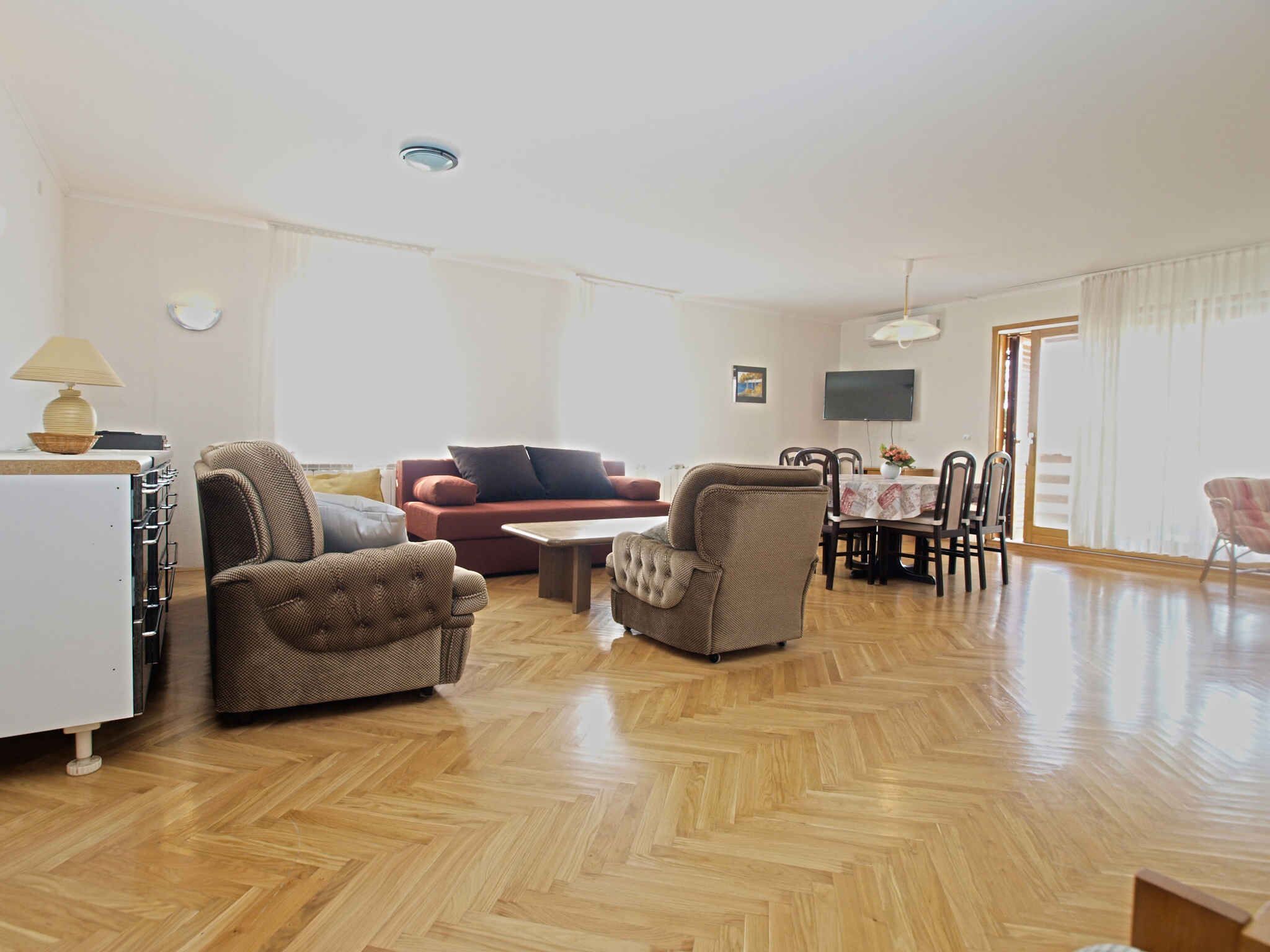 Ein großzügiger Wohnraum mit Holzdielen im Fischgrätmuster, einem roten Sofa, zwei Sesseln, einem Couchtisch und einem Essbereich mit Tisch und Stühlen.
