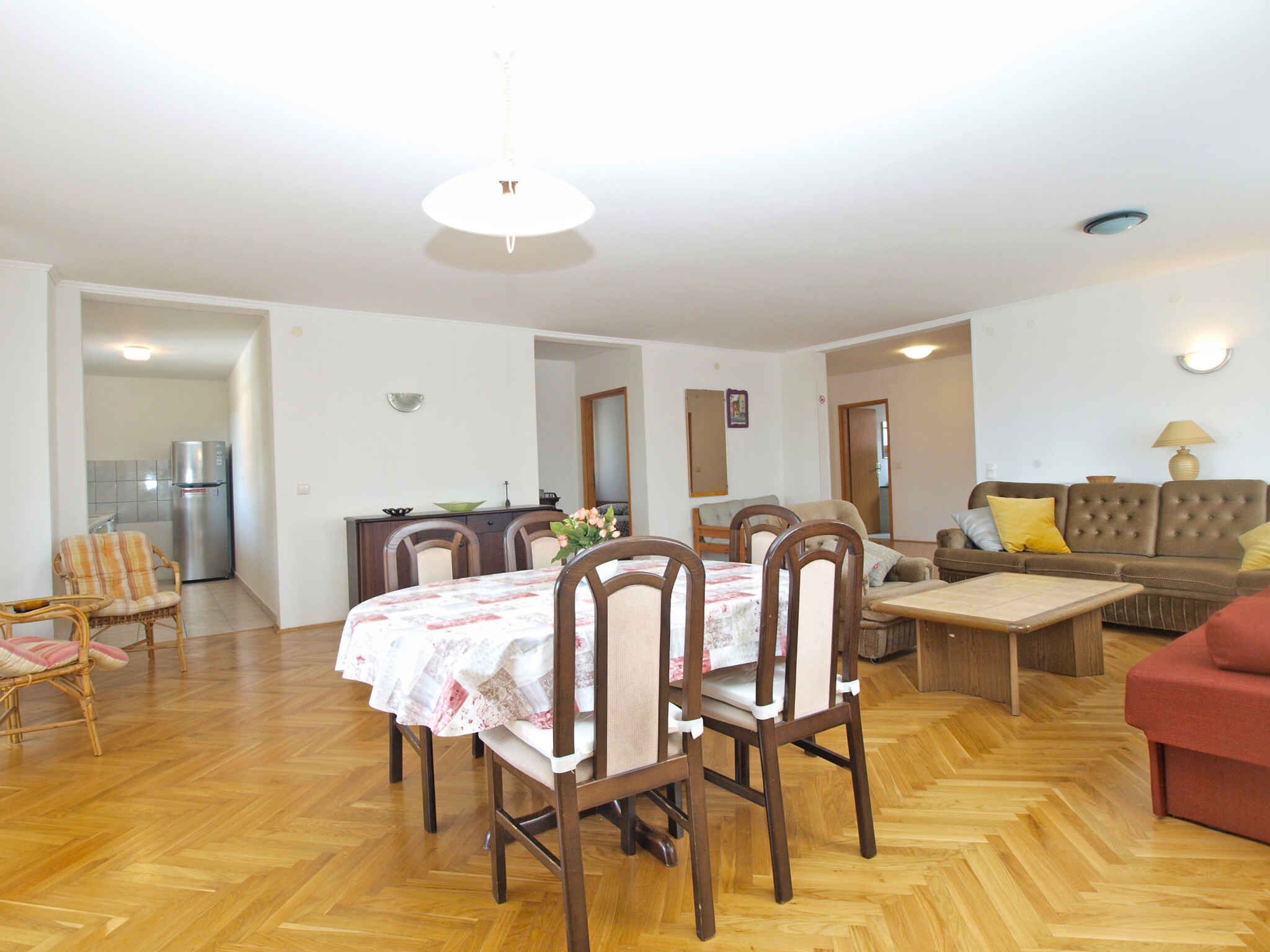 Uma sala ampla com uma mesa de jantar, cadeiras, um sofá e um sofá vermelho. É visível uma cozinha ao fundo.