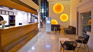 A modern hotel lobby with a reception desk, seating area, and a staircase leading to an upper level.
