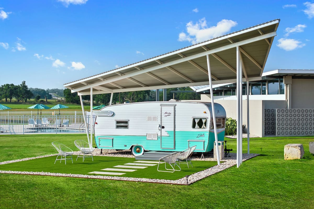 Una caravana antigua estacionada debajo de un pabellón moderno, con un césped y mobiliario exterior.