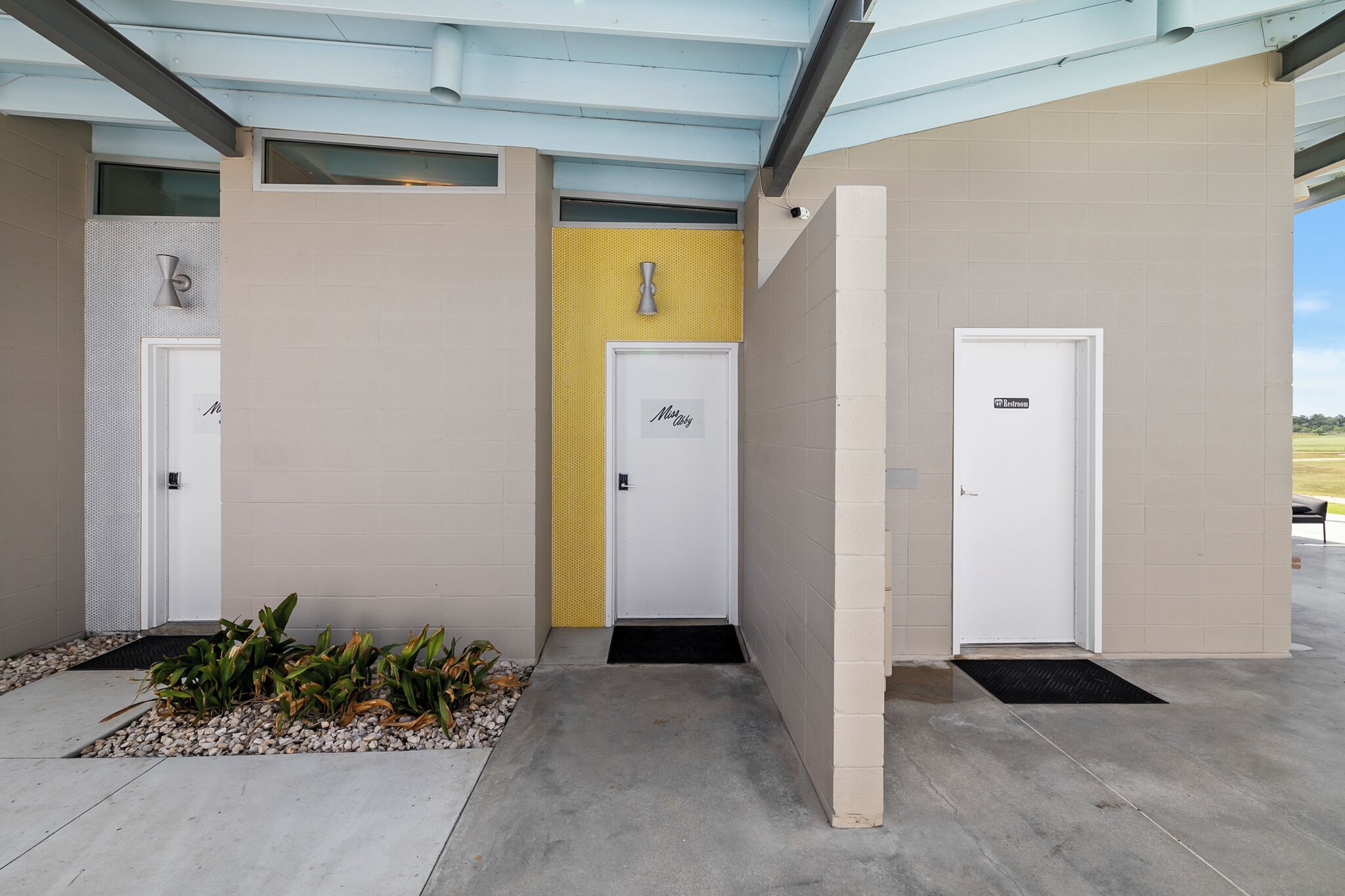 Une façade d’immeuble avec trois portes, chacune ornée d’un logo différent, et un petit jardin agrémenté de plantes et de galets.