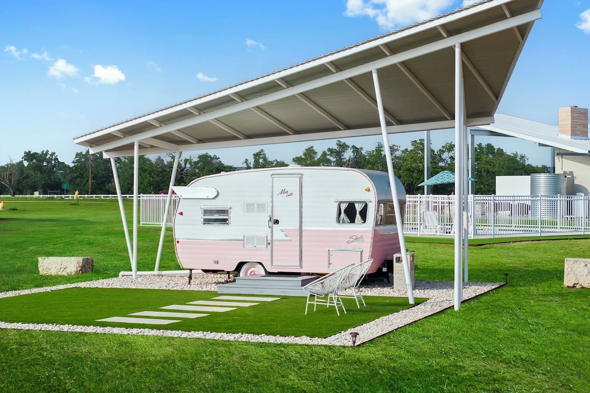 Una caravana rosa estacionada bajo una estructura cubierta, con una cerca blanca y zonas verdes a su alrededor.