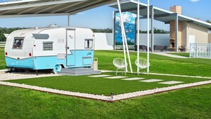 Un camping-car moderne est garé dans un espace verdoyant bien entretenu, avec un coin salon extérieur abrité et un bâtiment contemporain dont le mur du fond est décoré d’une fresque.