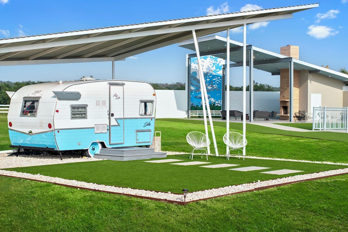 Una autocaravana moderna estacionada en un césped bien cuidado, con un área de estar cubierta y un edificio contemporáneo con un mural de fondo.