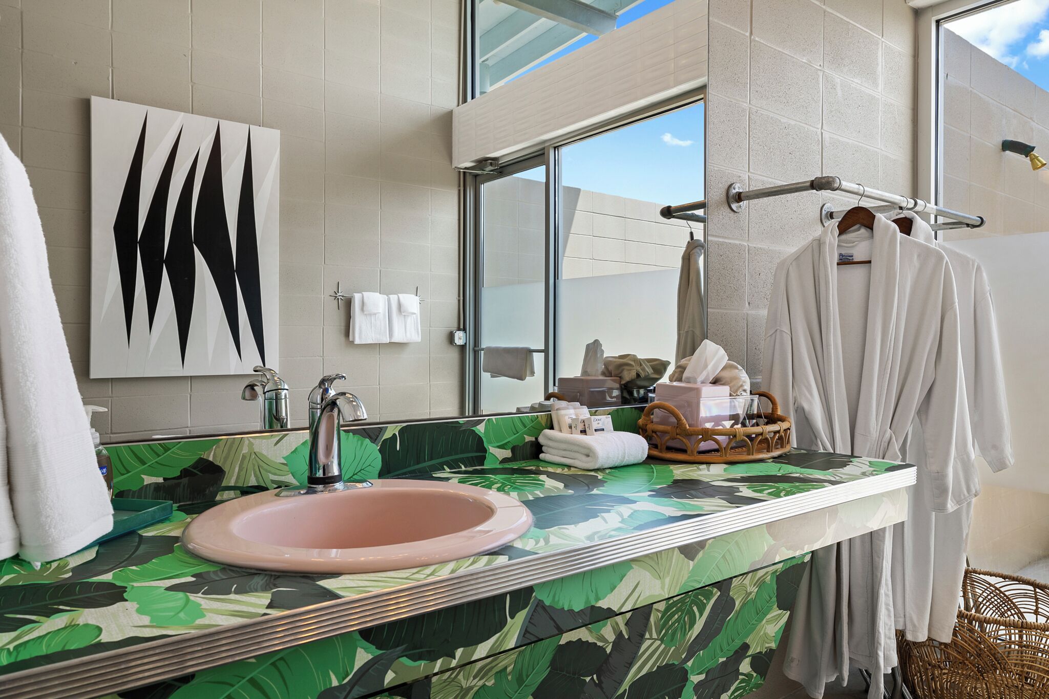 Une salle de bain avec un grand miroir, un lavabo et une fenêtre. On y trouve des serviettes et un panier contenant des articles de toilette, posés sur le comptoir.