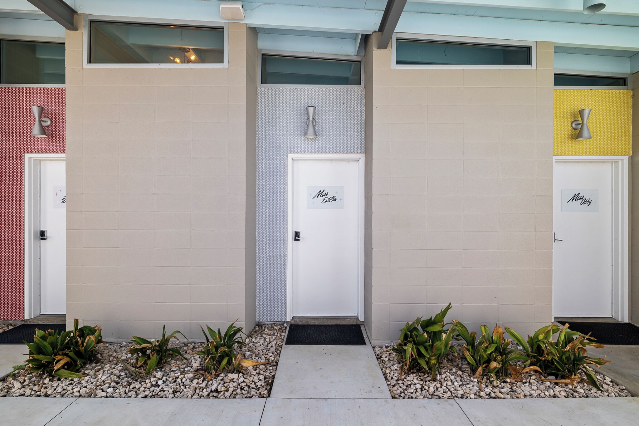 Une rangée de trois portes, chacune portant l’inscription « Mademoiselle Estelle », est adossée à un mur de couleur beige, devant lequel se trouve un petit jardin.