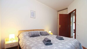 A neatly made bed with a floral-patterned bedspread and two pillows, a bedside lamp, and a small nightstand with a lamp. - Holiday apartment with air conditioning (Pješćana Uvala)