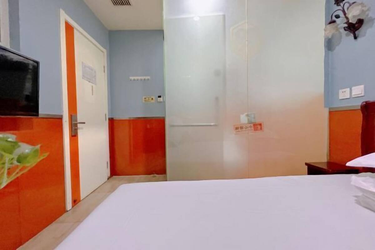 Quarto de hotel com cama, televisão, porta e luminária fixada na parede.