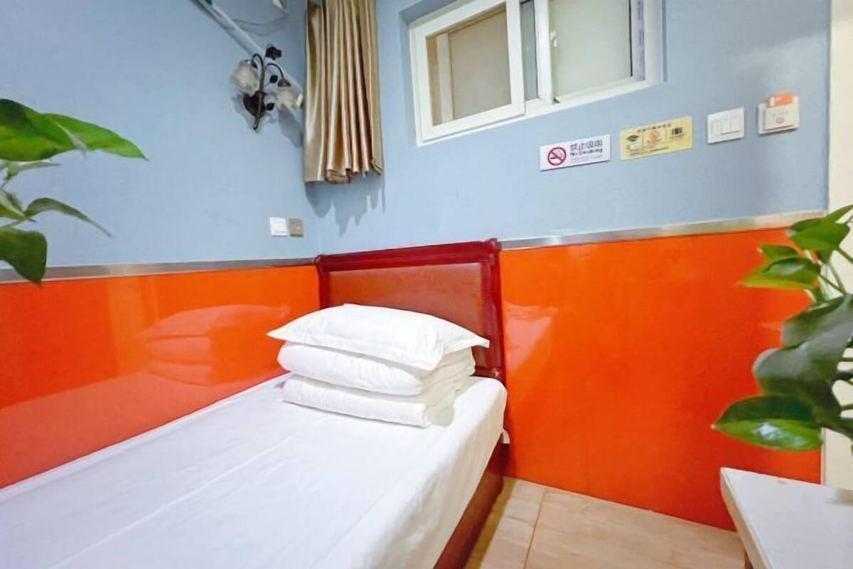 Quarto pequeno, com uma cama de solteiro, paredes laranja e azuis, uma janela com cortinas e uma placa na parede.