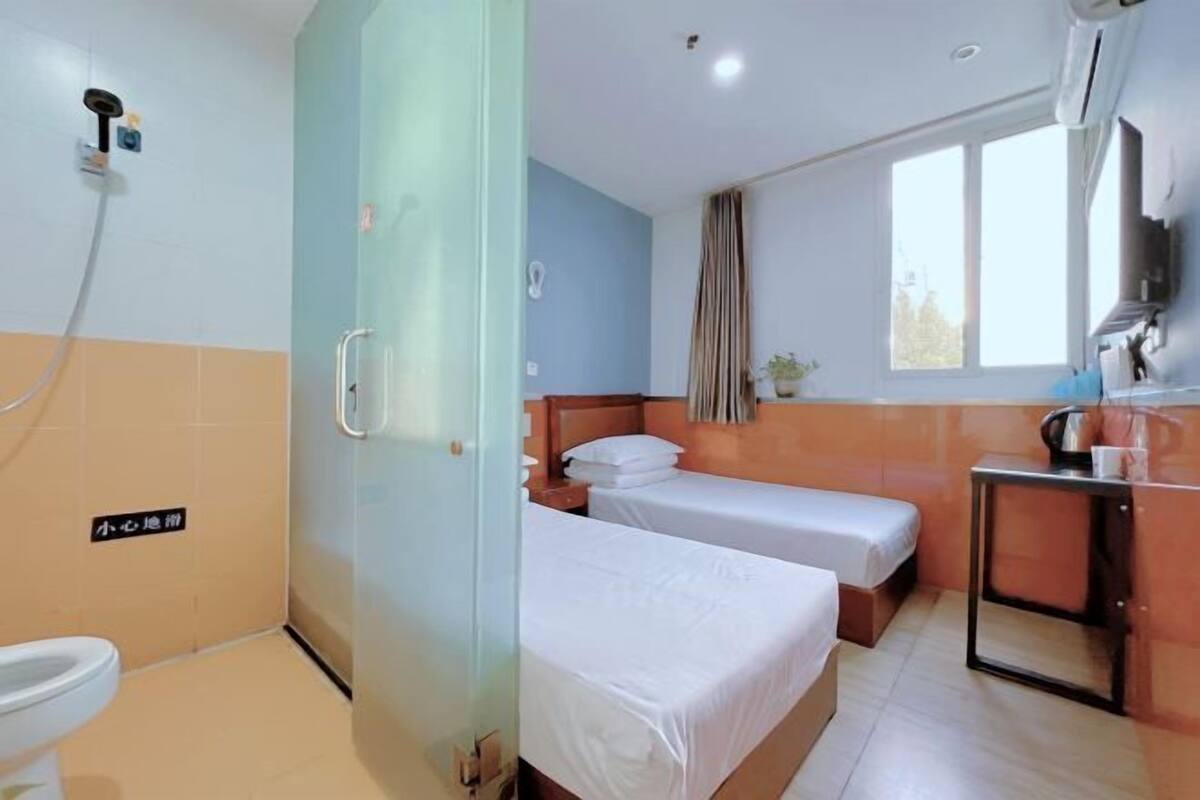 Quarto de hotel com duas camas individuais, banheiro com chuveiro, uma mesinha com chaleira e janela com cortinas.