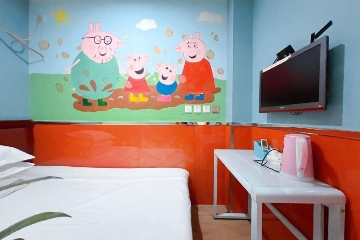 Quarto de hotel com cama, televisão presa na parede, uma mesinha com chaleira e um mural colorido na parede.