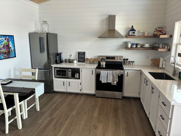 A modern kitchen with white cabinetry, a stainless steel refrigerator, a microwave, an oven, and a sink. There is a wooden dining table with chairs, a painting on the wall, and a sign above the stove. - Red Jellybean: great ocean views and sunsets close to town (Twillingate)