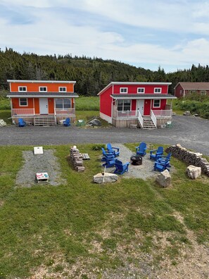Two red buildings with white railings, a gravel area with blue chairs, and a forested hill in the background. - Red Jellybean: great ocean views and sunsets close to town (Twillingate)