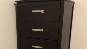 Un archivador negro con varios cajones y pomos metálicos, situado frente a una pared lisa con suelo de madera.