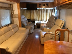 Un interior de caravana confortable con un sofá, dos sillones, una mesita y un televisor montado en la pared.
