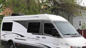 Valkoinen pakettiauto, jonka kyljessä on teksti »Vista Cruiser», on pysäköity sorapintaiselle alueelle.