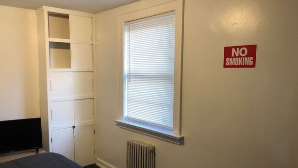 A bedroom with a bed, a radiator, a window with blinds, a white wardrobe, and a 'NO SMOKING' sign on the wall.