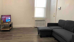 A living room with a dark gray sectional sofa, a small wooden table with a TV, and a window with blinds.