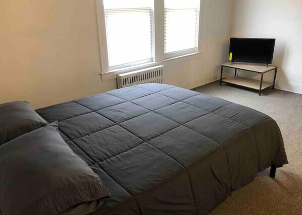 A bedroom with a bed, a radiator, a television on a stand, and a window with blinds.