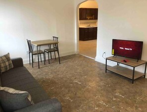 Uma sala de estar com sofá, mesa de jantar com cadeiras, TV em um suporte e uma cozinha visível por um arco.